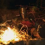 Guerrier médiéval casqué, assis près d’un feu dans une forêt sombre